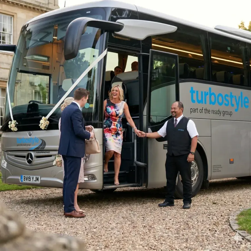 A professional Turbostyle driver in a black waistcoat assists a well-dressed female wedding guest as she disembarks from a silver luxury Mercedes-Benz coach on a gravel driveway.