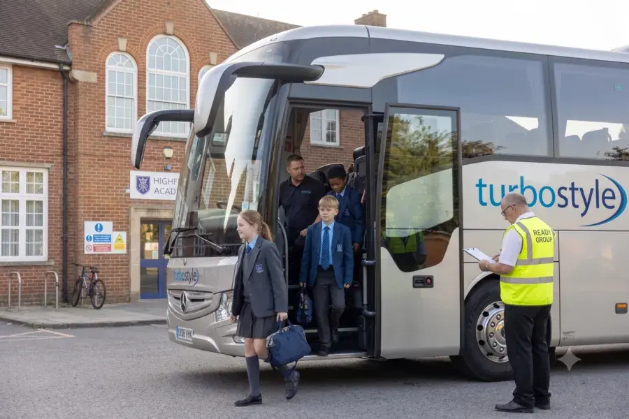 Side view of students in school uniform stepping down from the coach door onto the pavement next to a high-visibility staff supervisor.