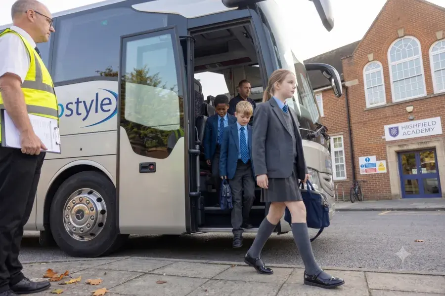 Low-angle shot of students exiting the silver Turbostyle coach outside Highfield Academy while a staff member holds a clipboard.