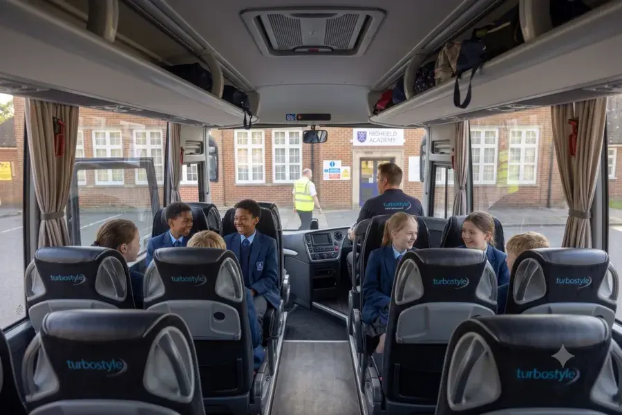 Interior view looking down the center aisle of the coach toward the front windshield, showing students seated and Highfield Academy visible outside.